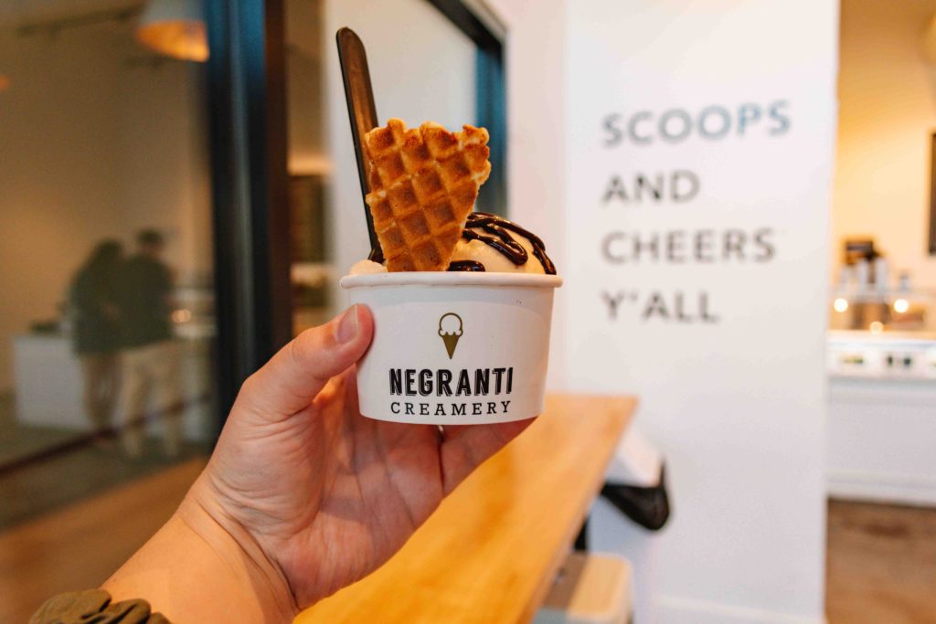 outstretched hand holding a scoop of sheep's milk ice cream in a cup with a waffle cone chip in it and a sign in the back that reads "Scoops and cheers y'all"