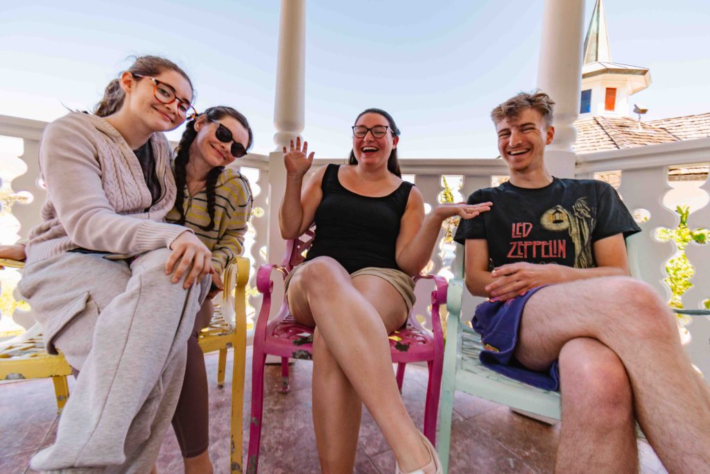 Four adults cousins (3 girls, 1 boy) sitting on colorful pastel chairs under a gazebo