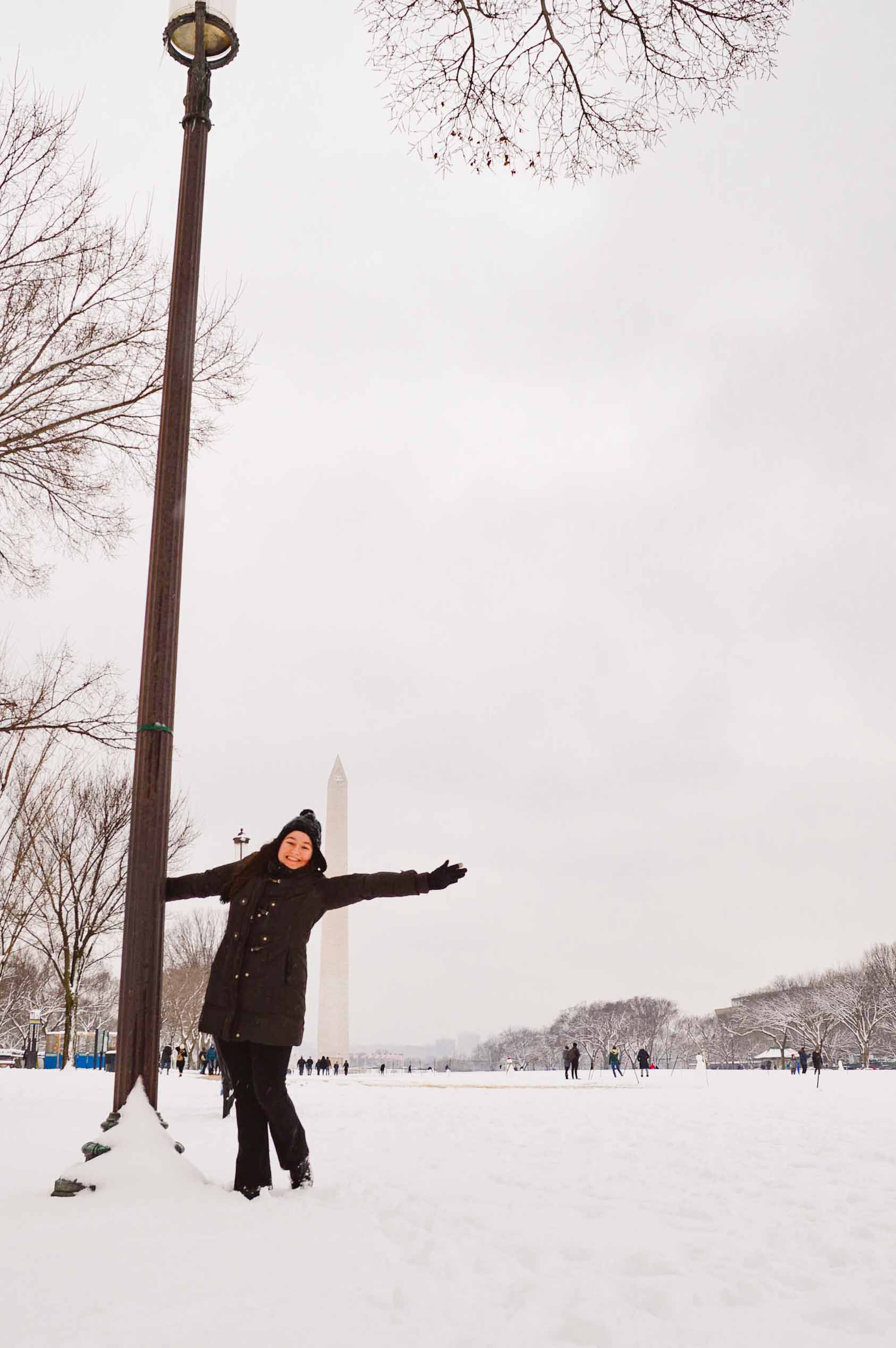 woman hanging off a light post during a snow storm with the Washington Monument behind her