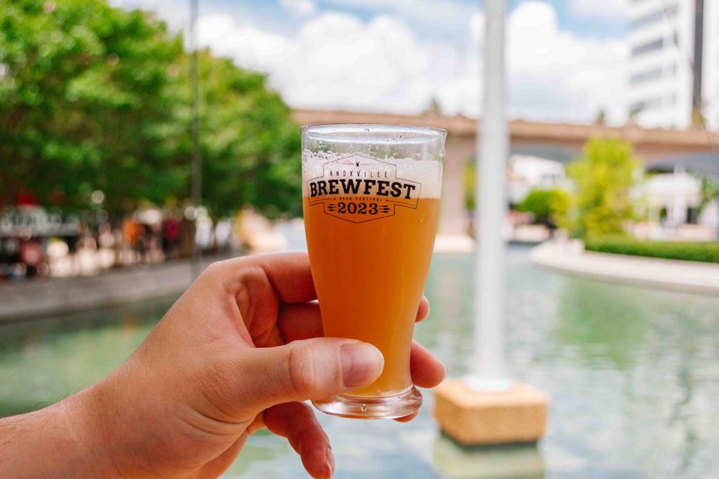 woman's outstretched hand holding a 5oz pour of beer in a plastic cup that says BrewFest