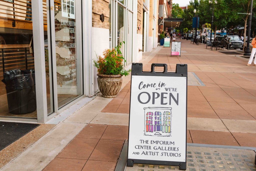 View of art gallery sign welcoming people in with two other gallery's signs in the distance along the same sidewalk.