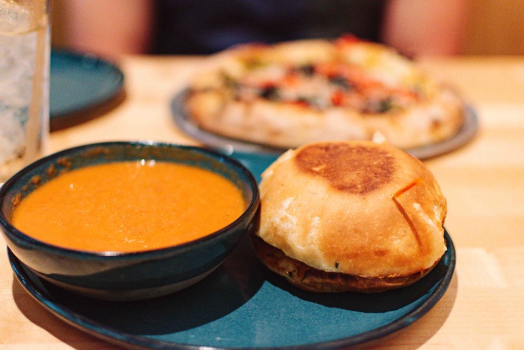 Small sandwich and tomato chipotle soup with pizza in the background.