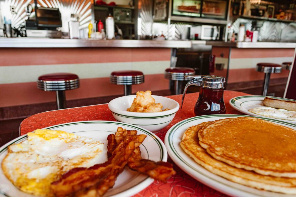 Classic breakfast in a 1950's stainless steel diner including pancakes, bacon, eggs, and hashbrowns with a syrup jug behind.