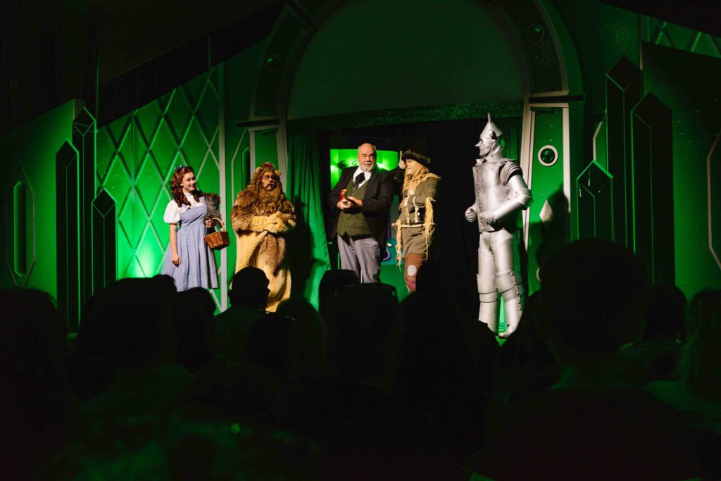 Dorothy, Scarecrow, Tin Man, and Cowardly Lion standing with the Wizard on a green stage