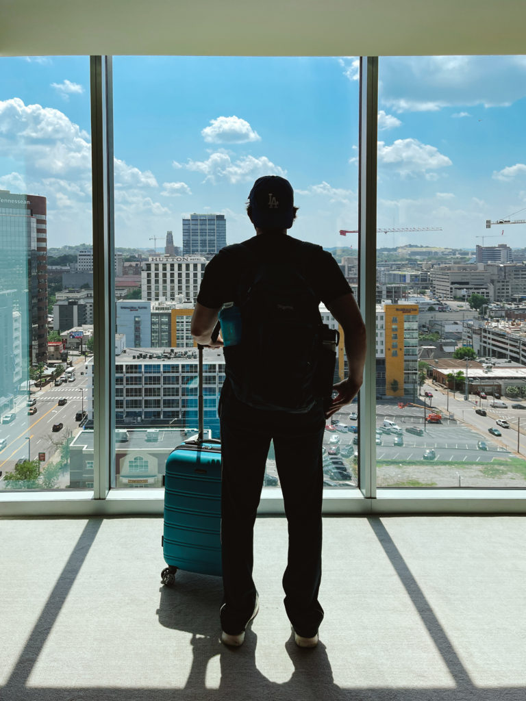Man standing with his suitcase and a backpack on looking out a floor to ceiling window at the Nashville cityscape