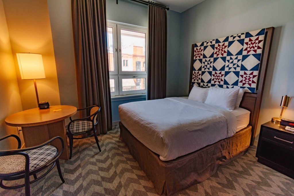 View of a hotel room interior with queen bed, table and two chairs, and tall window