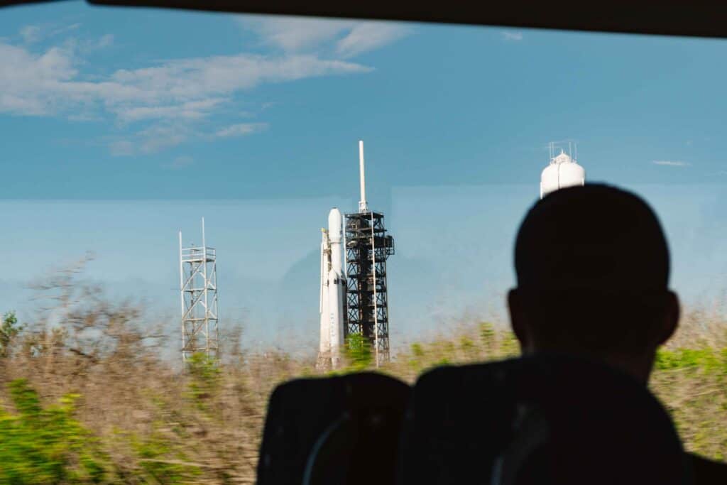Bus tour of Kennedy Space Center behind the gates where a man in silhouette is looking out the window at the Nancy Grace Roman telescope on the rocket ready for launch.