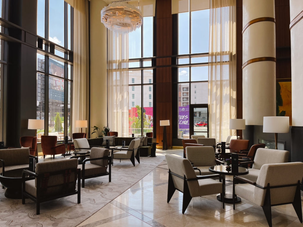 Collections of lounge chairs and tables in an open hotel lobby with large floor to ceiling windows and large chandelier