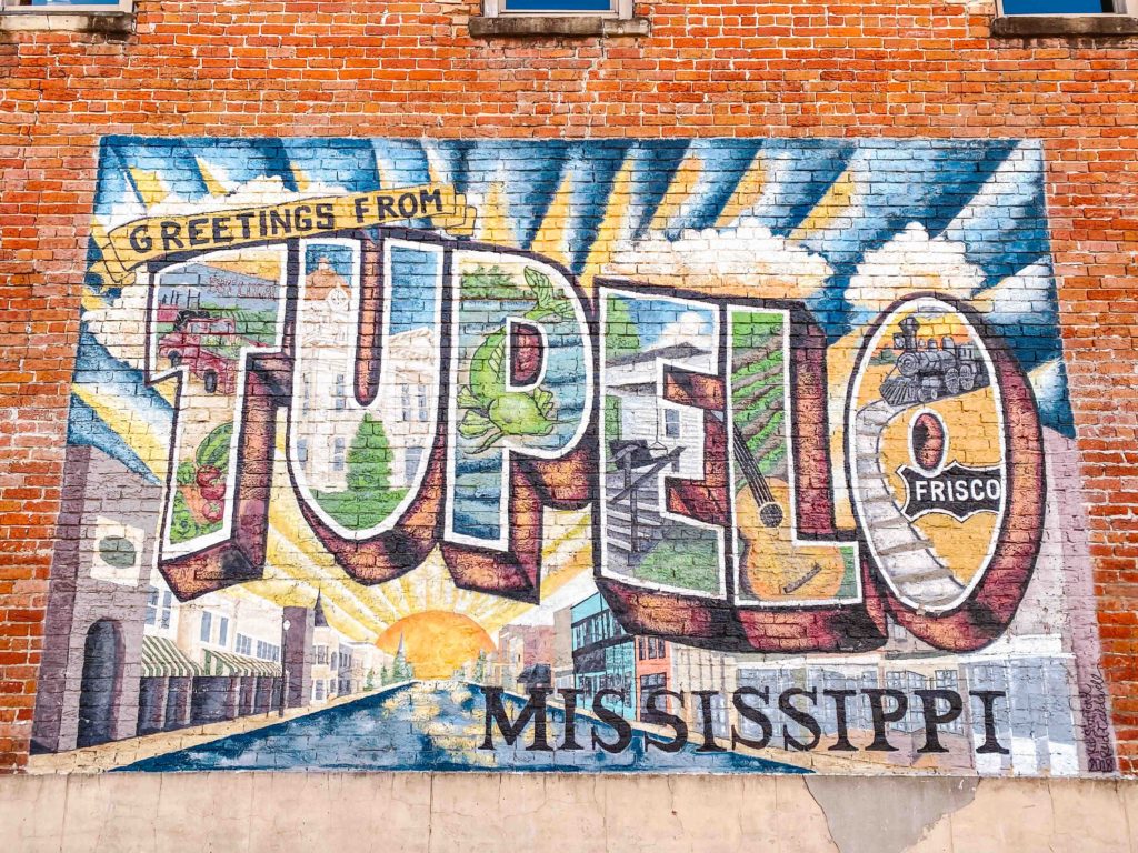 Mural that says Greetings from Tupelo Mississippi that looks like a brightly colored postcard on a brick wall