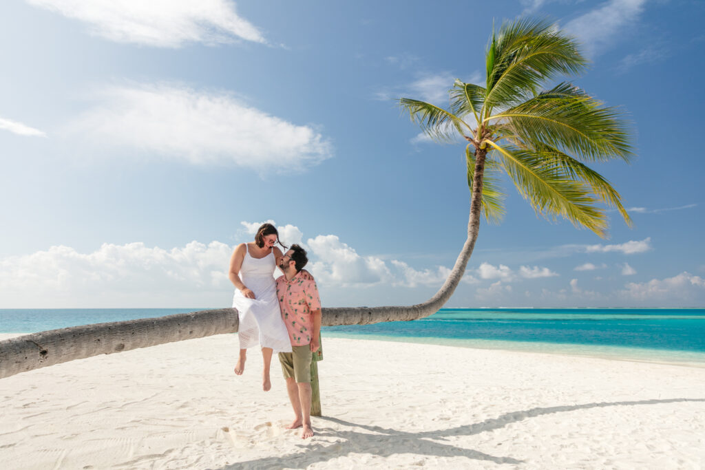 Man and woman on their honeymoon, a photoshoot with a horizontal palm tree on the beach at Conrad Maldives with turquoise water behind