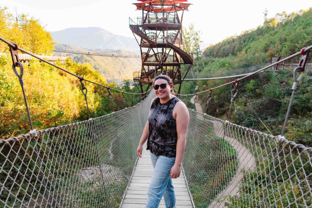 Woman standing on a suspension bridge leading to an observation tower with a long sky bridge in the background and green rolling mountains of Gatlinburg Tennessee