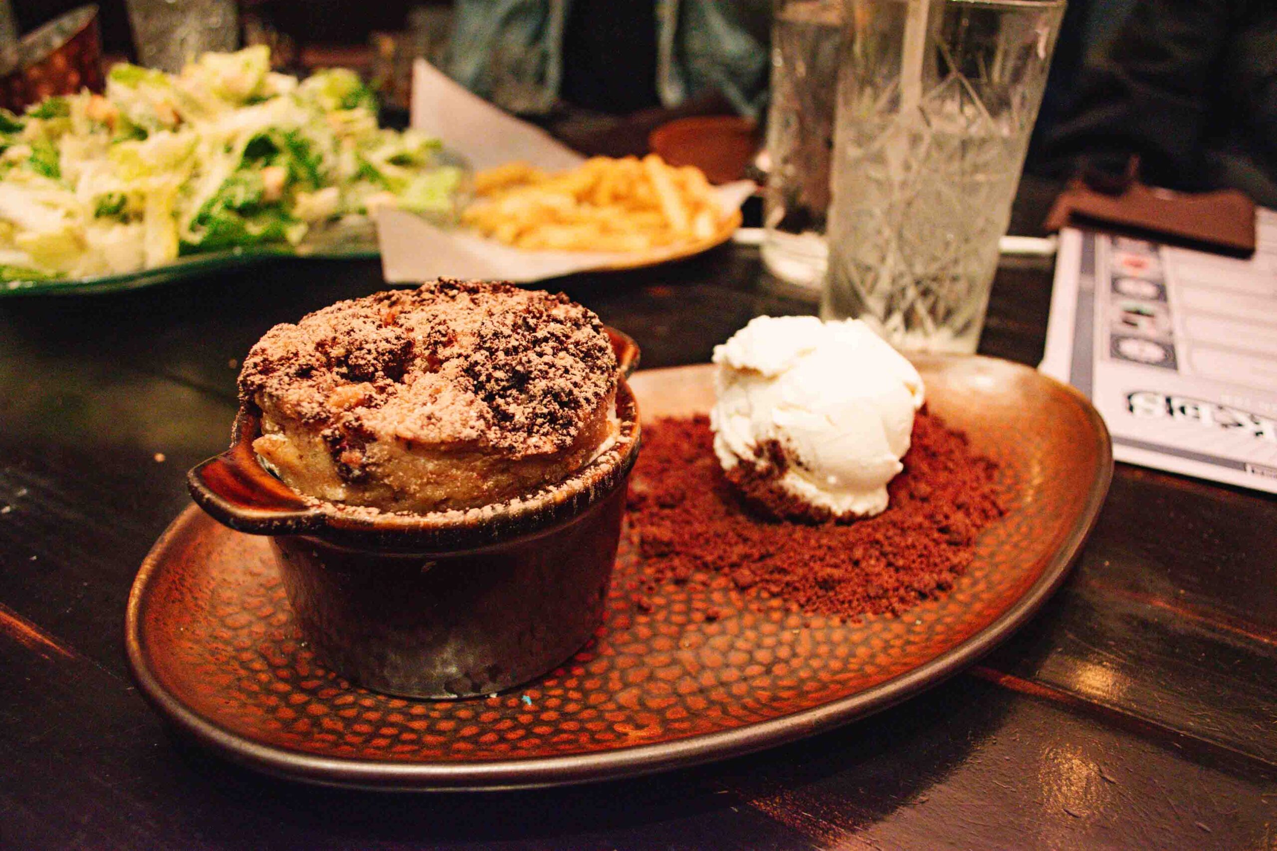Triple chocolate bread pudding baked in a ramekin and served on a brown plate with a scoop of vanilla ice cream to the side on top of cookie crumbs.
