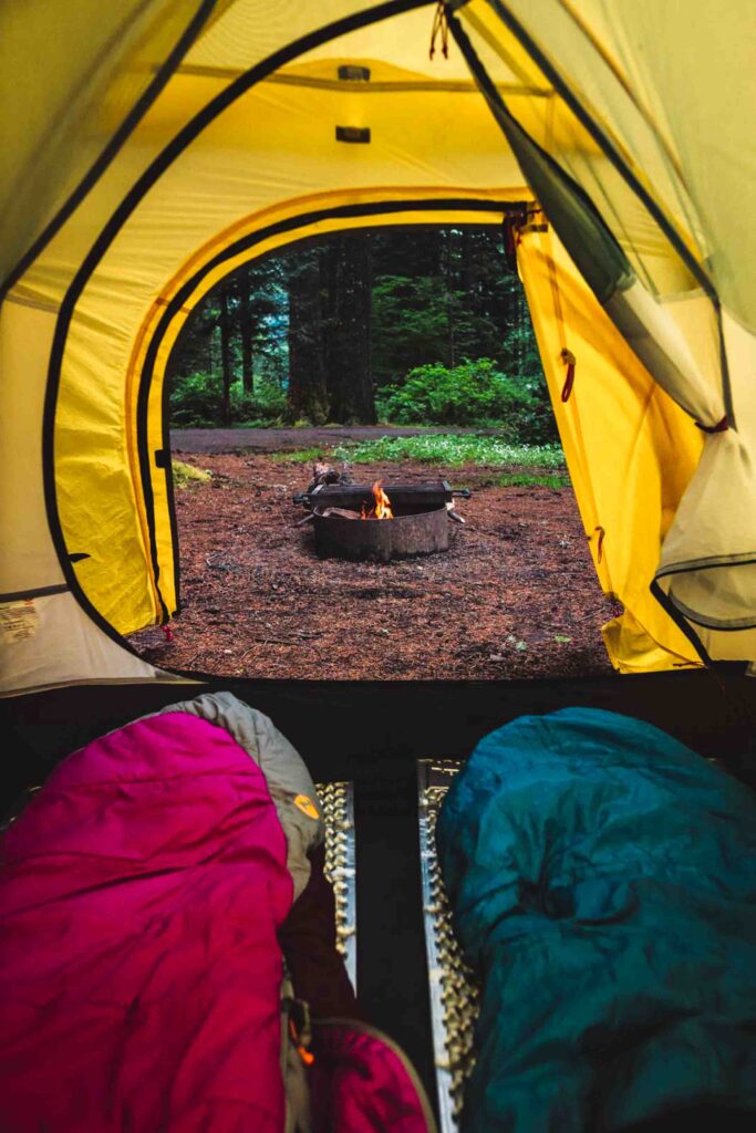 View looking out of a yellow tent with two sleeping bags at the foot of the tent and fire going in a fire pit centered on the tent's opening