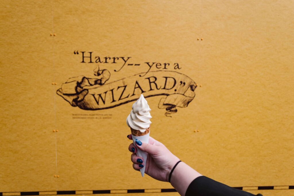 Woman holding a cone of butterbeer ice cream in front of a yellow wall with a quote that reads "Harry...yer a wizard"