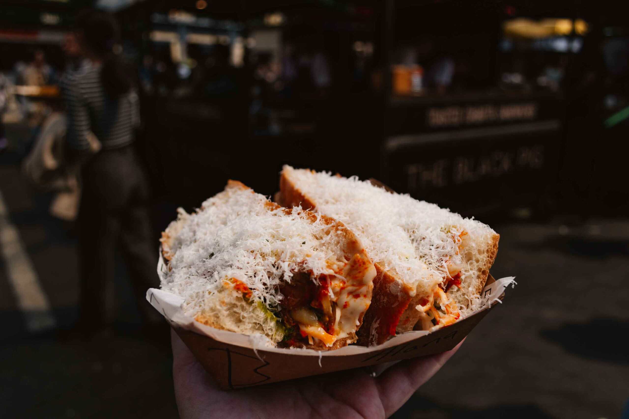 Woman's outstretched hand holding a pork sandwich topped with finely shredded parmesan from Borough Market in london