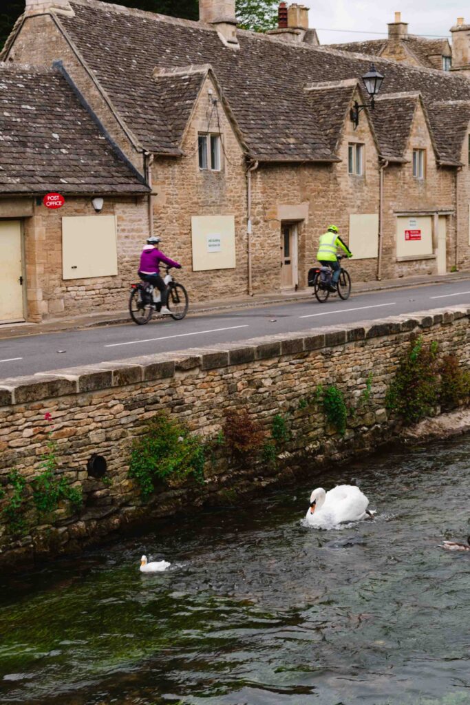 View of bikers passing through Bibury along the water that geese are swimming in