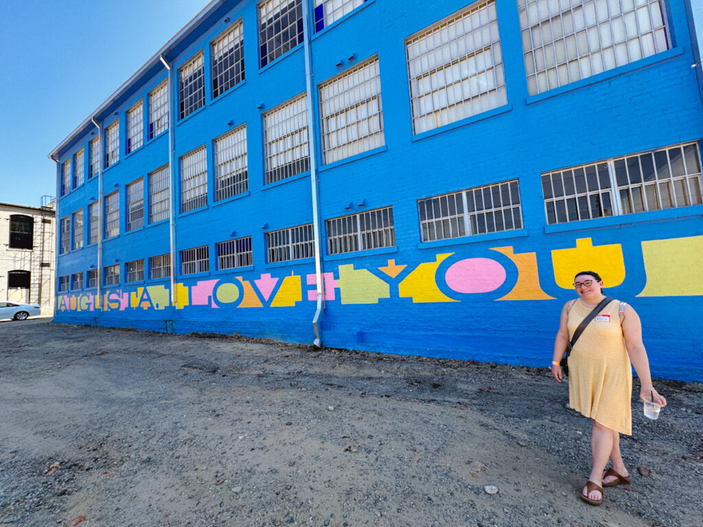 Augusta I Love You mural with woman in yellow dress standing in front