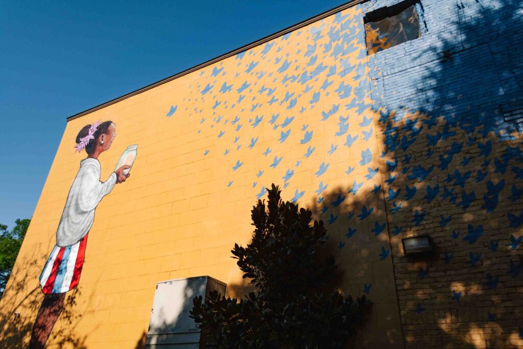 Mural of a young Black girl releasing blue butterflies that make up the right portion of the wall, which has been painted blue
