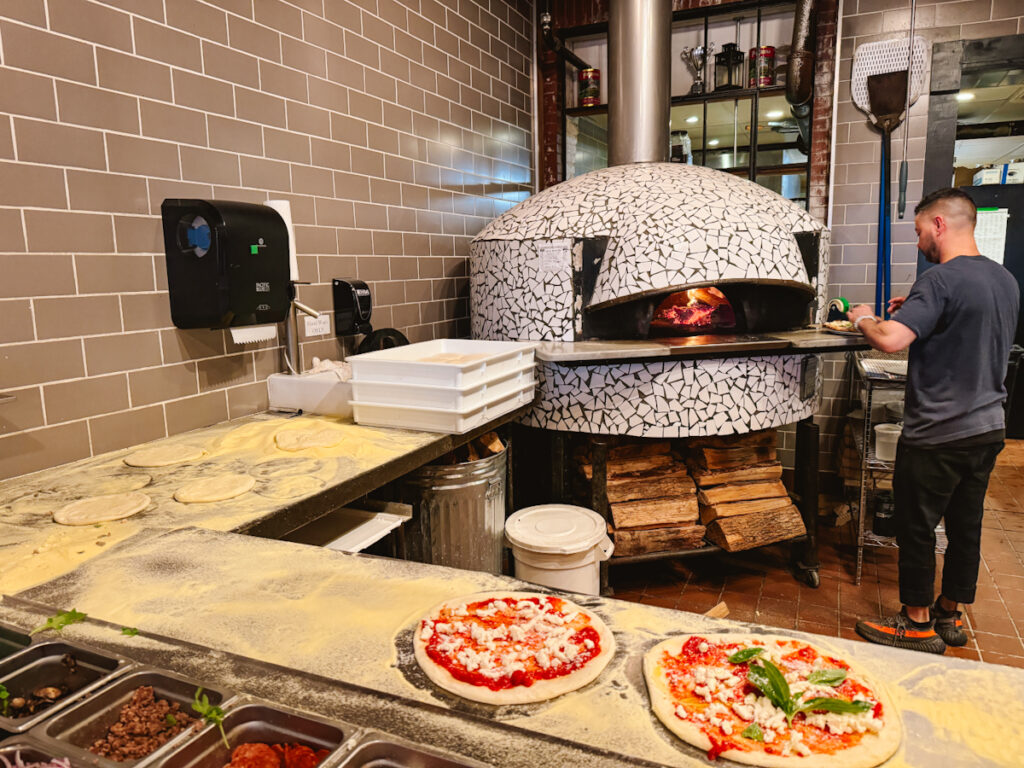 View of an open kitchen in a restaurant with a woodfired pizza oven in the back, a must try restaurant in Salem