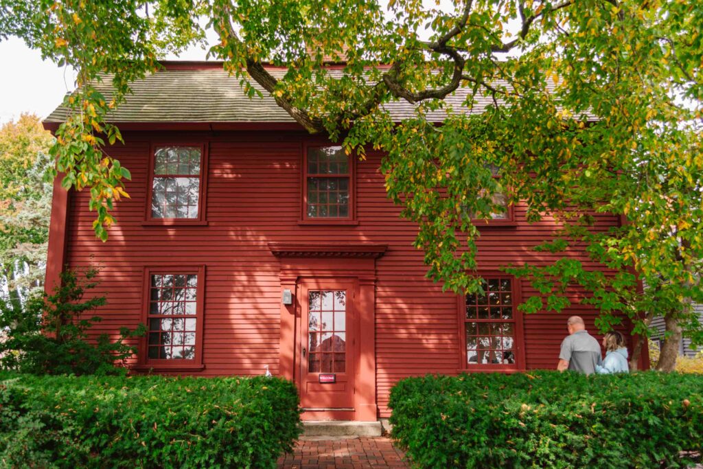 Exterior of the home that was the birthplace of Nathaniel Hawthorne with lush green tree overhead, two bushes to the right and left, and a red painted exterior