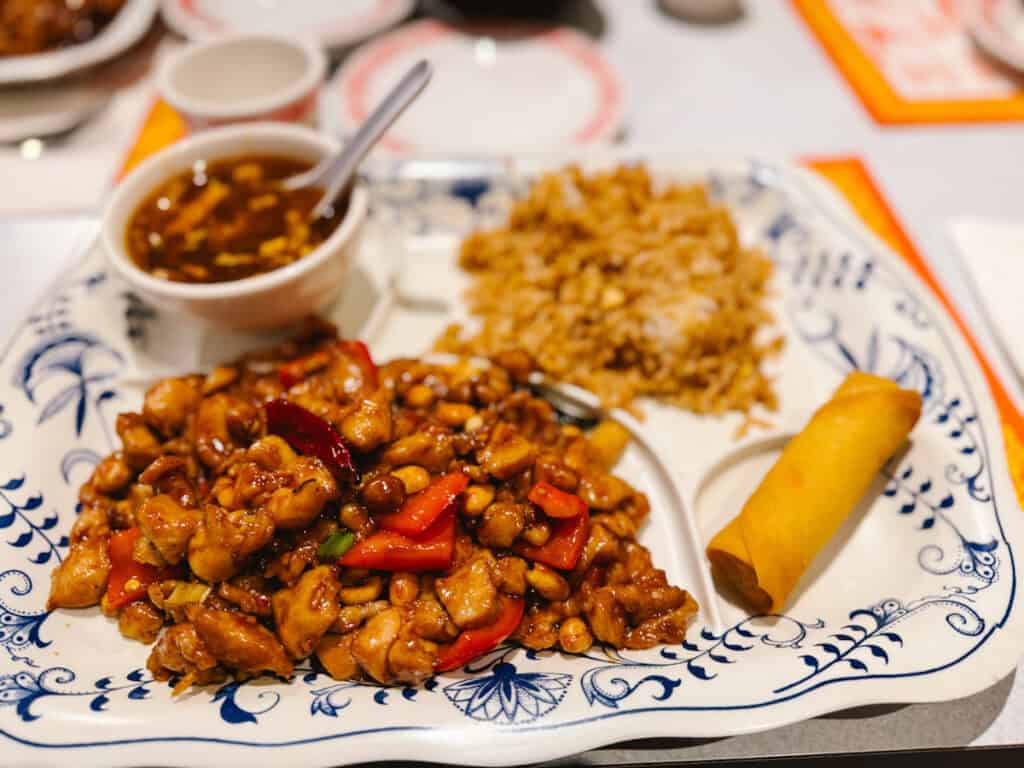 Schezuan Chinese food on a lunch platter with kung pao chicken, a vegetable roll, brown rice, and soup to the top left.