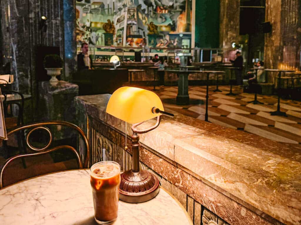 Iced mocha on a marble table with library lamp to the side in an art deco coffee hall with intricate mural on the back wall and tiled floor