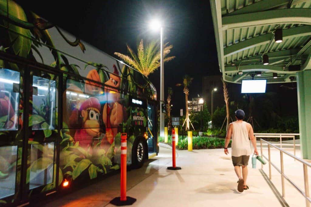 Man walking to shuttle bus to his left with Donkey Kong and Diddy Kong on the exterior of the bus, at night with a sign that reads Stella Nova, indicating where the shuttle is going