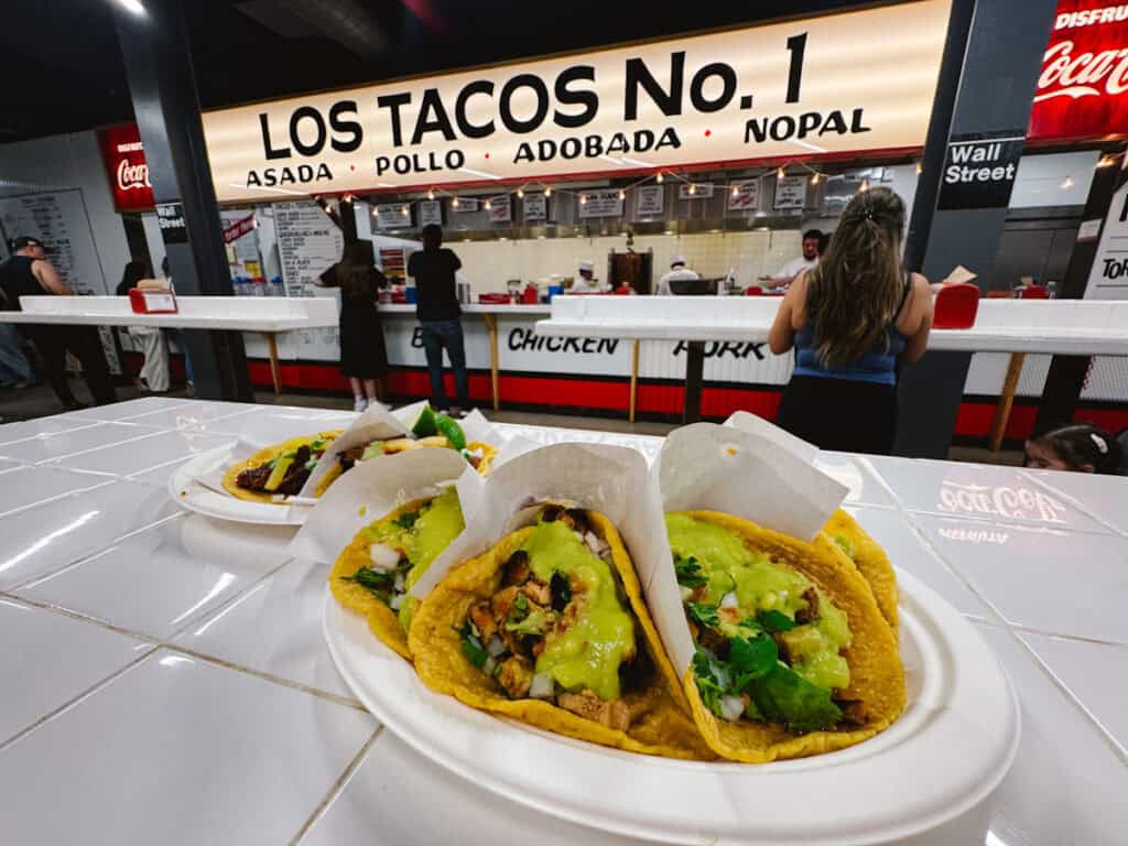 Three tacos on a disposable plate from Los Tacos No. 1 with the restaurant name sign in the background