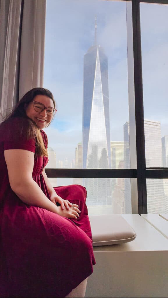 Woman sitting on a windowsill in New York city hotel downtown with a view of the One World Trade Center