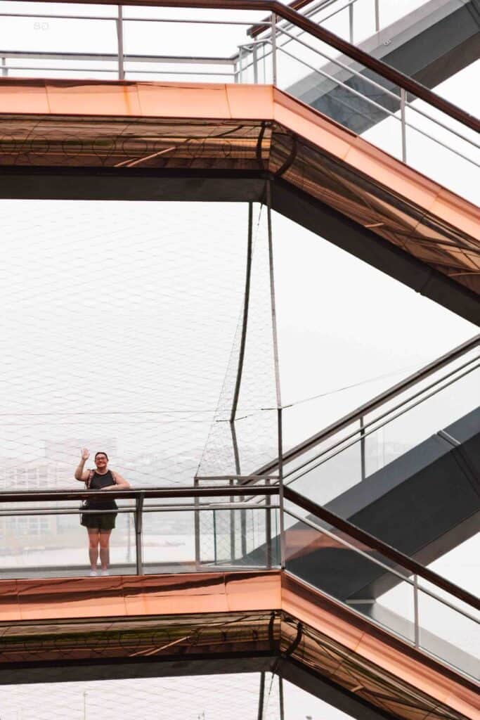 Woman standing a good distance from the camera and waving, while standing on a funky geometric pattern building called the Vessel in NYC