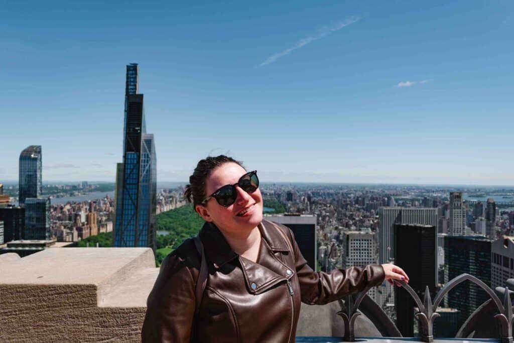 Woman smiling and tilting her head toward the camera while wearing a brown leather jacket with an unobstructed view of NYC behind her facing Central Park to the north