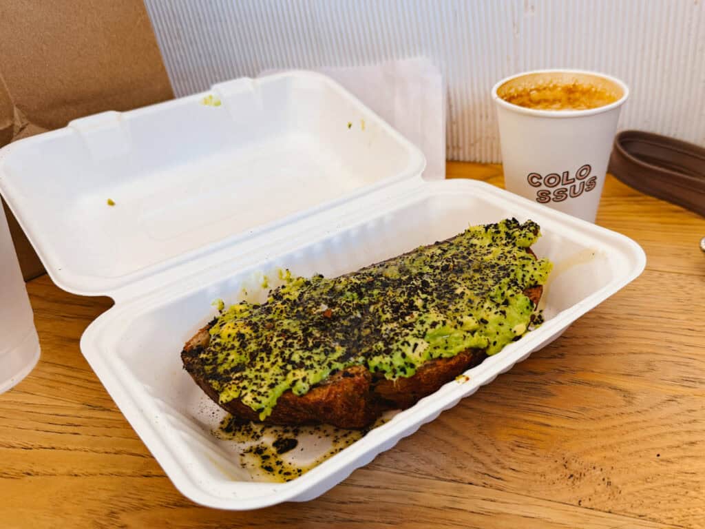 To-go box with thick avocado toast topped with black sea salt and coffee behind to the right, where the cup reads "Colossus"