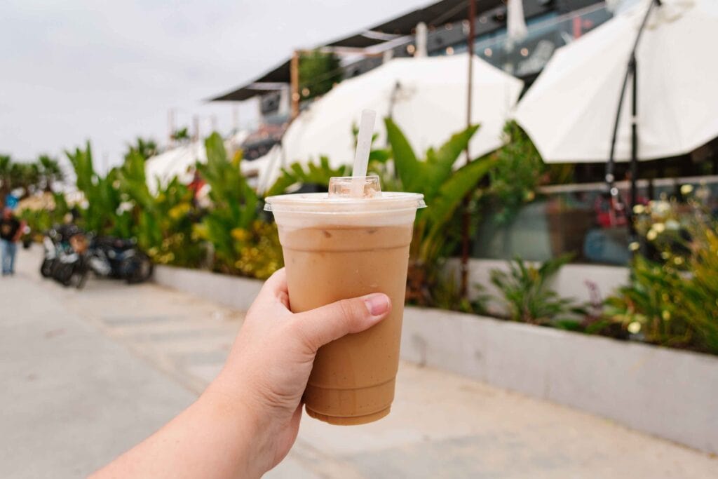 Woman's outstretched hand holding an iced coffee outside a tropical restaurant with big leafy bushes in front