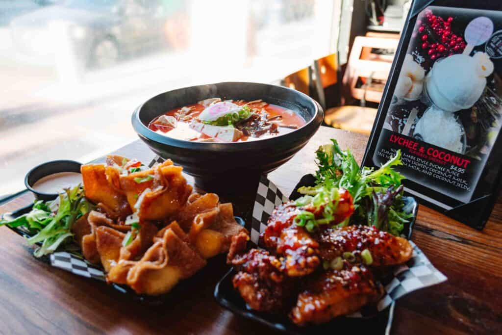 Table of food including udon ramen in a big black bowl, and two tapas of Japanese comfort food including cream cheese wontons and wings