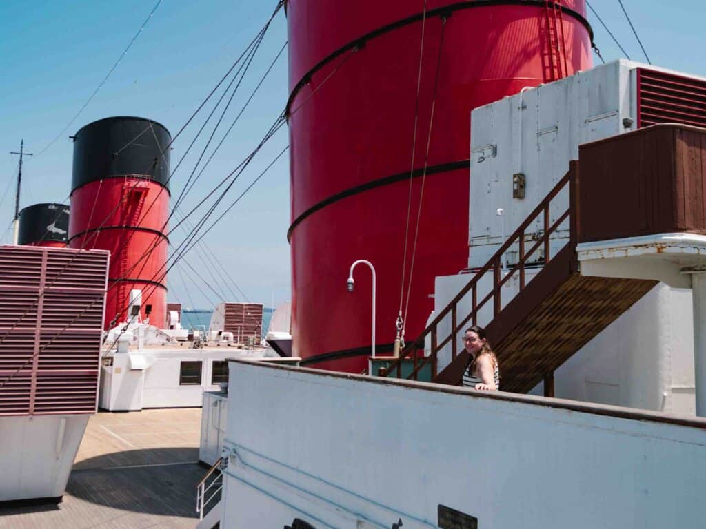 Woman standing on the Queen Mary's uppermost deck with big red exhaust towers along the ship's deck