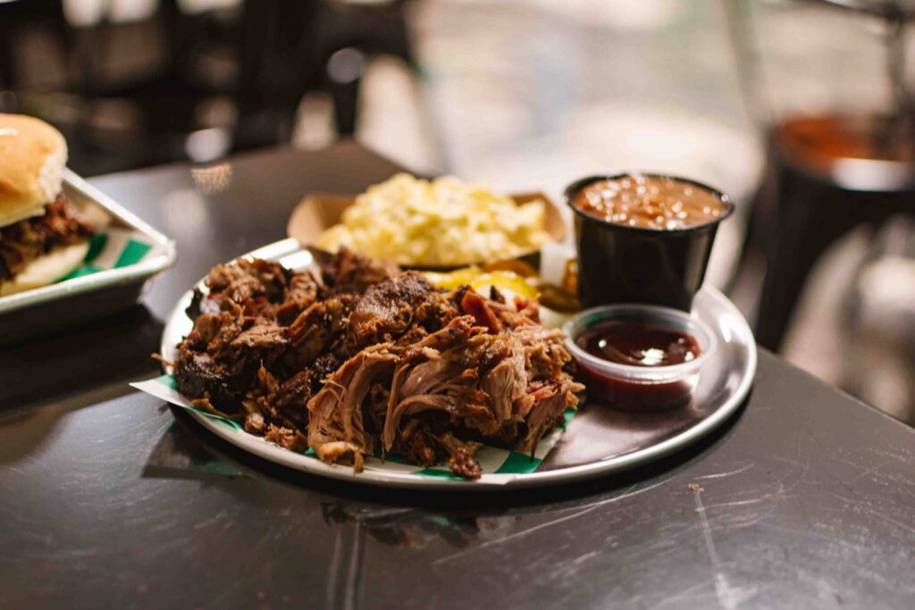 Plate of BBQ food with pulled pork, brisket, and sides of baked beans in a small cup and potato salad