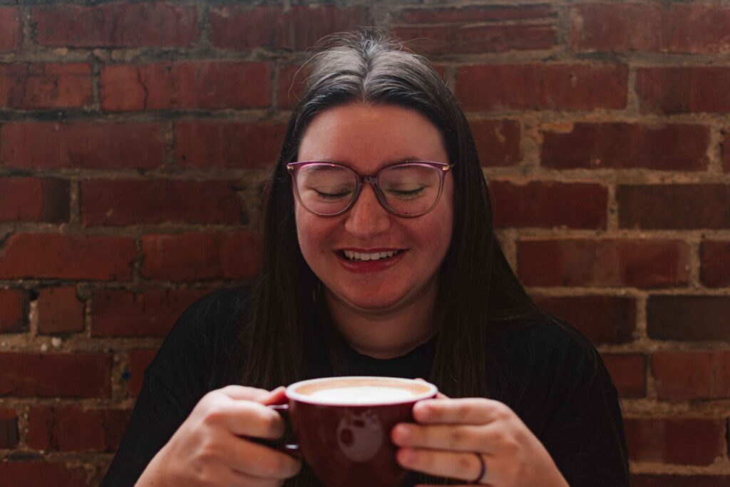 Woman enjoying a cup of coffee, she's smiling looking down at the mug she's holding with a brick wall behind her