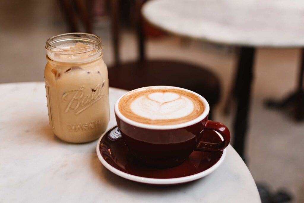 An iced coffee in a Mason jar and a hot mocha in a burgundy mug and saucer in front on a white marble round table at a coffee shop in Columbia Tennessee called Hank's Coffee Station