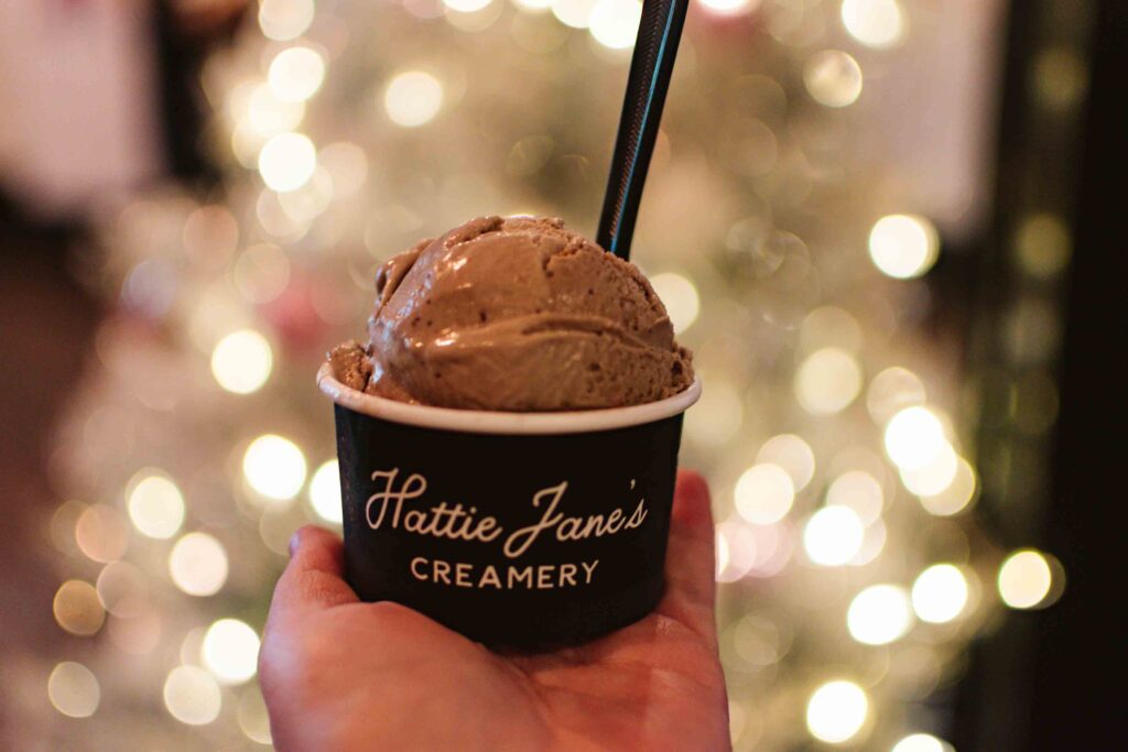 Woman's outstretched hand holding a cup of chocolate ice cream with a black plastic spoon stuck inside to the right with a white Christmas tree and lights in the background