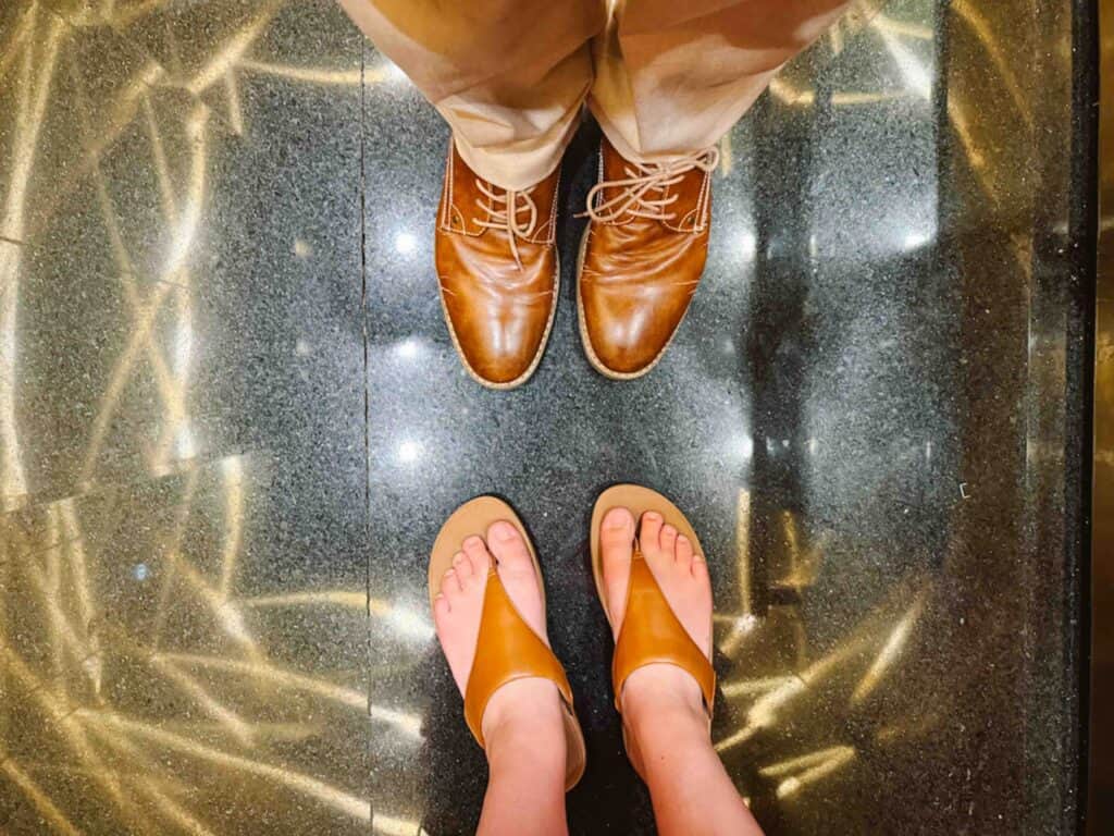 Looking down at two pairs of shoes in an elevator where the man has brown dress shoes and the woman is wearing brown formal sandals