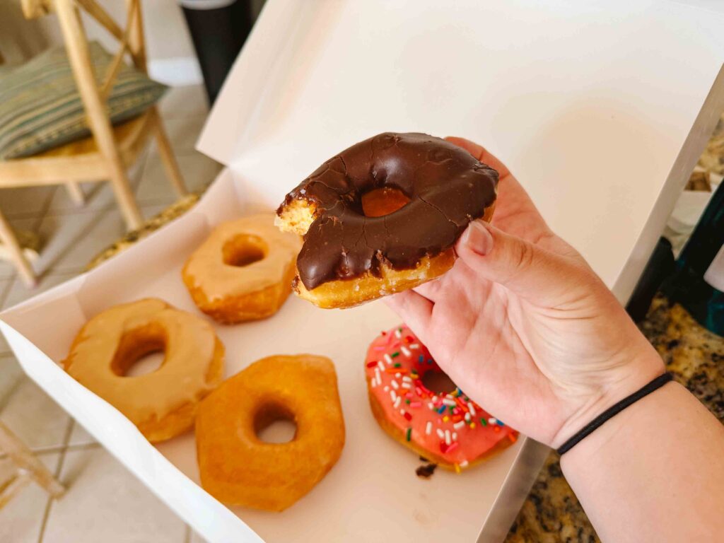 Holding out a chocolate covered donut over a box of 6 miscellaneous donuts