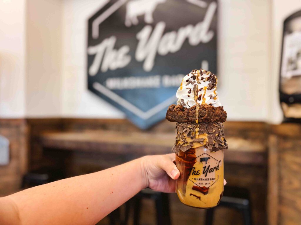Woman holding a crazy milkshake that is peanut butter chocolate brownie with The Yard ice cream shop logo on the wall behind her hand