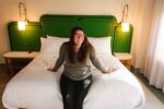 The Waymark Hotel room interior with woman in comfy clothes sitting on the foot edge of the bed and smiling