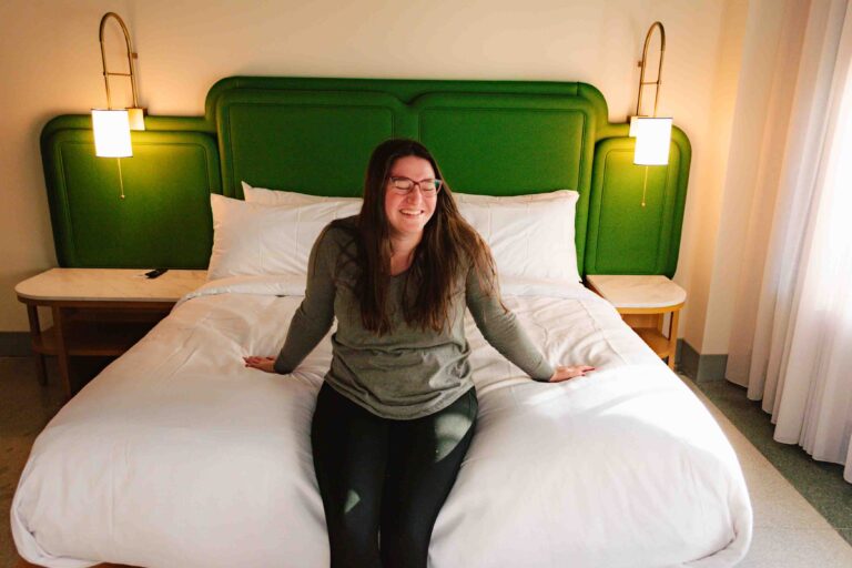 The Waymark Hotel room interior with woman in comfy clothes sitting on the foot edge of the bed and smiling