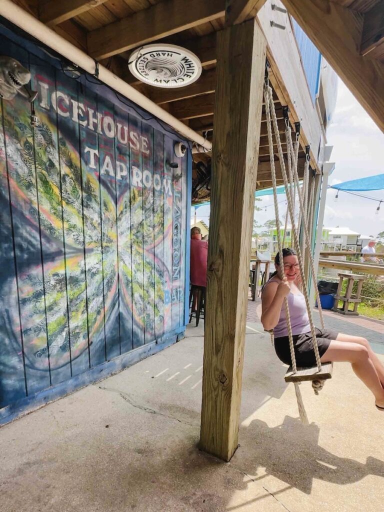 Woman on a swing smiling at the camera from the side with a mural on a building to the left behind her that says Icehouse Taproom, the bar she is at.