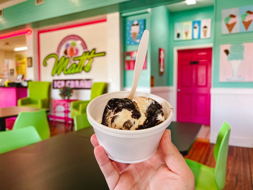 Woman's outstretched hand holding a styrofoam bowl of vanilla ice cream with hot fudge with a green and pink ice cream shop interior in the background
